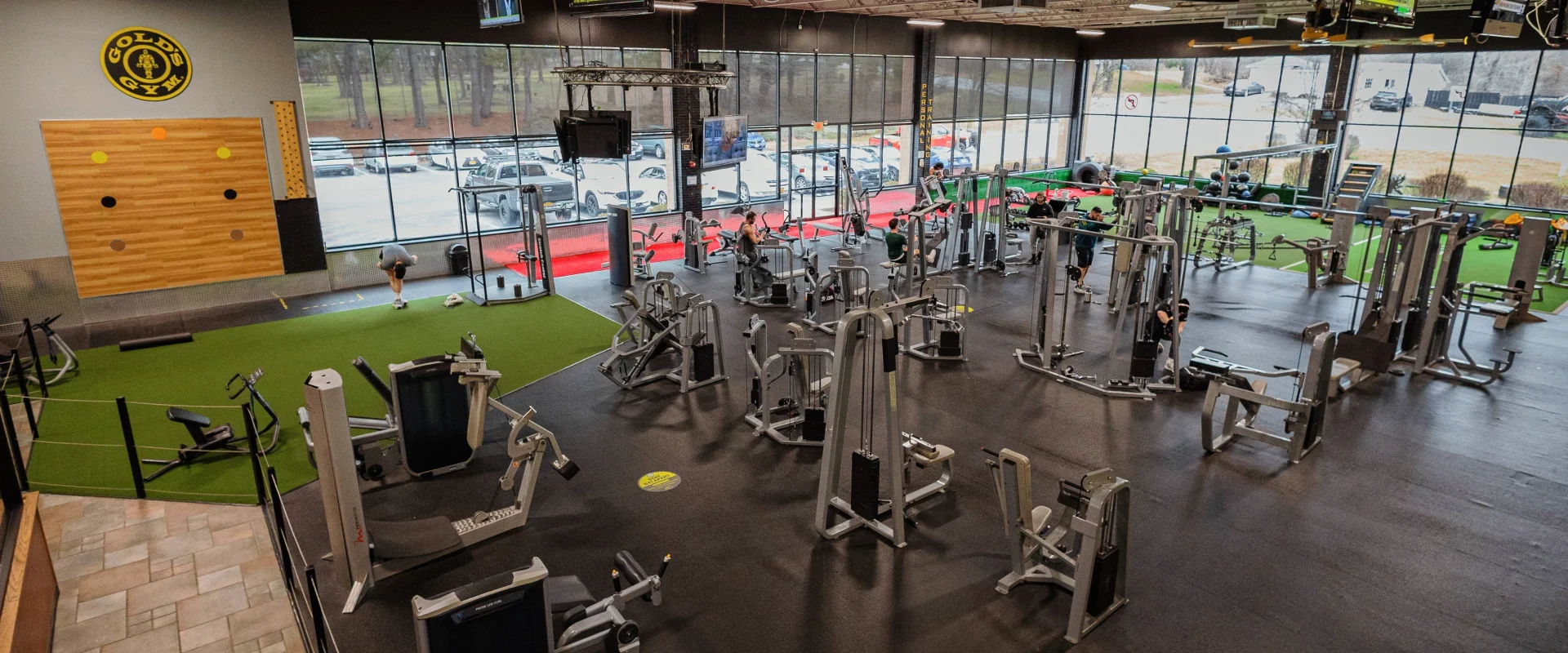 Gold's Gym New York (Poughkeepsie) gym floor showcasing a spacious layout with resistance machines, functional training turf, and a dedicated stretching area. Large windows flood the space with natural light, while multiple flat-screen TVs hang from the ceiling. The turf section features sleds, kettlebells, and functional equipment, with a climbing wall accenting the back wall near the Gold’s Gym logo.