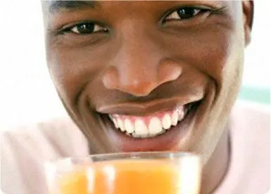 A smiling person with a bright, joyful expression holding a glass of orange juice in the foreground, with a blurred background.