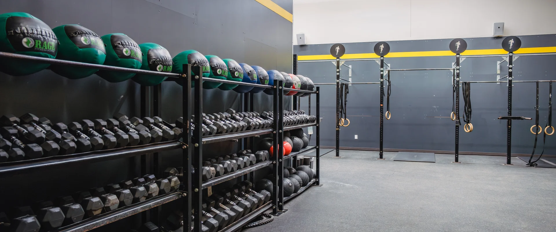 Gold’s Fit area in Austin (Westlake), featuring rows of green and black medicine balls, dumbbells neatly stacked on metal racks, and functional training equipment. The spacious setup includes wall-mounted rigs with gymnastic rings and pull-up bars, accented by a bold yellow stripe along the wall. The open floor space is ideal for dynamic, high-intensity workouts.