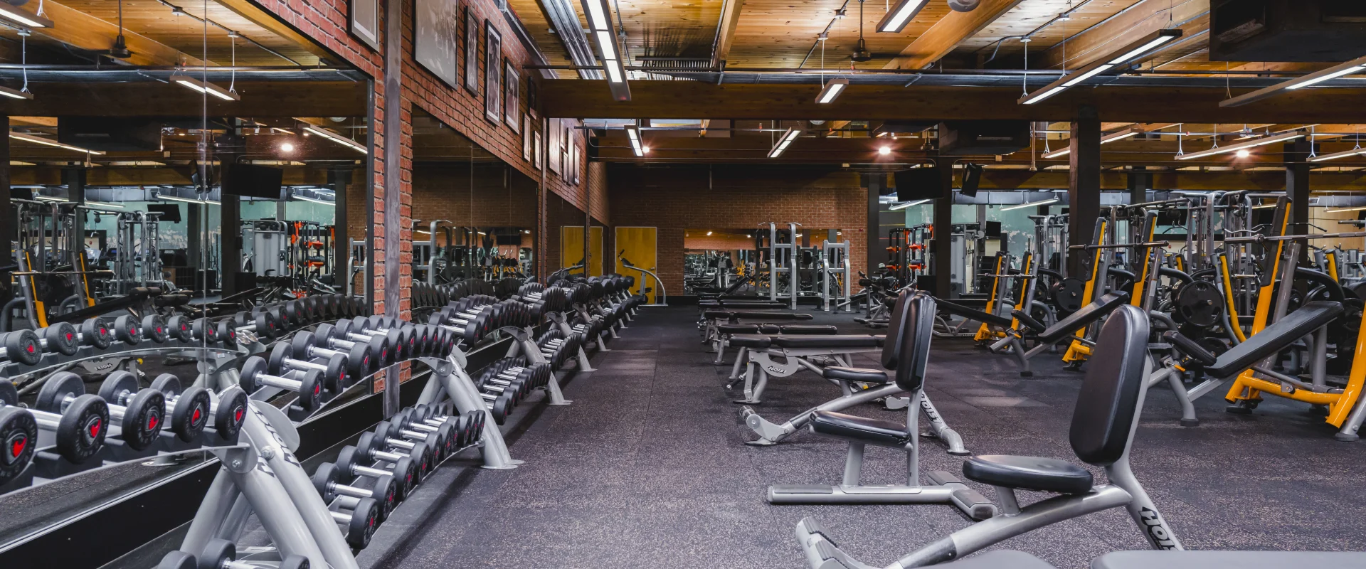 A gym area with rows of neatly organized dumbbells on racks, benches for workouts, mirrored walls, and a warm wooden ceiling with exposed beams.
