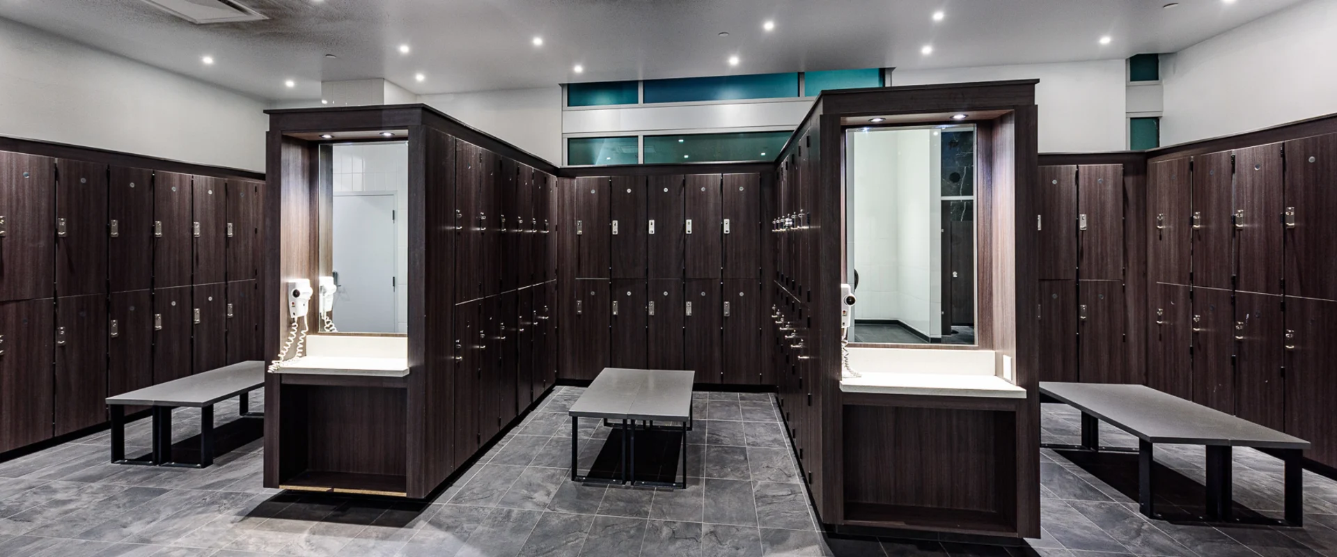 Locker room at Gold’s Gym Long Beach, featuring dark wood lockers, tiled floors, and built-in vanity stations with mirrors and hair dryers. Benches provide seating throughout the space, offering a functional and modern area for members to store belongings and freshen up. Recessed lighting enhances the sleek and organized design.