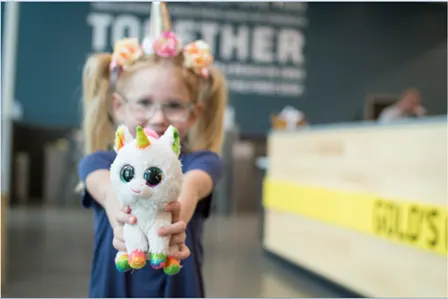 A young girl with a colorful flower crown holds a plush unicorn toy in the foreground, with a "Together" sign visible in the background.