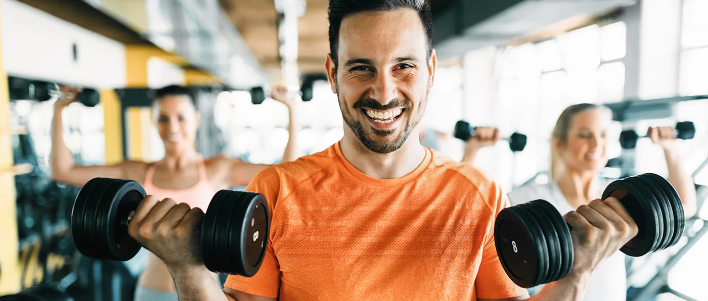 A smiling man in an orange shirt is lifting weights in a gym, with other people visible in the background.