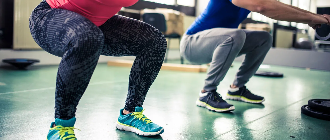 The image shows the lower bodies of two people wearing athletic clothing and sneakers, standing on a green floor in what appears to be a gym or fitness studio.