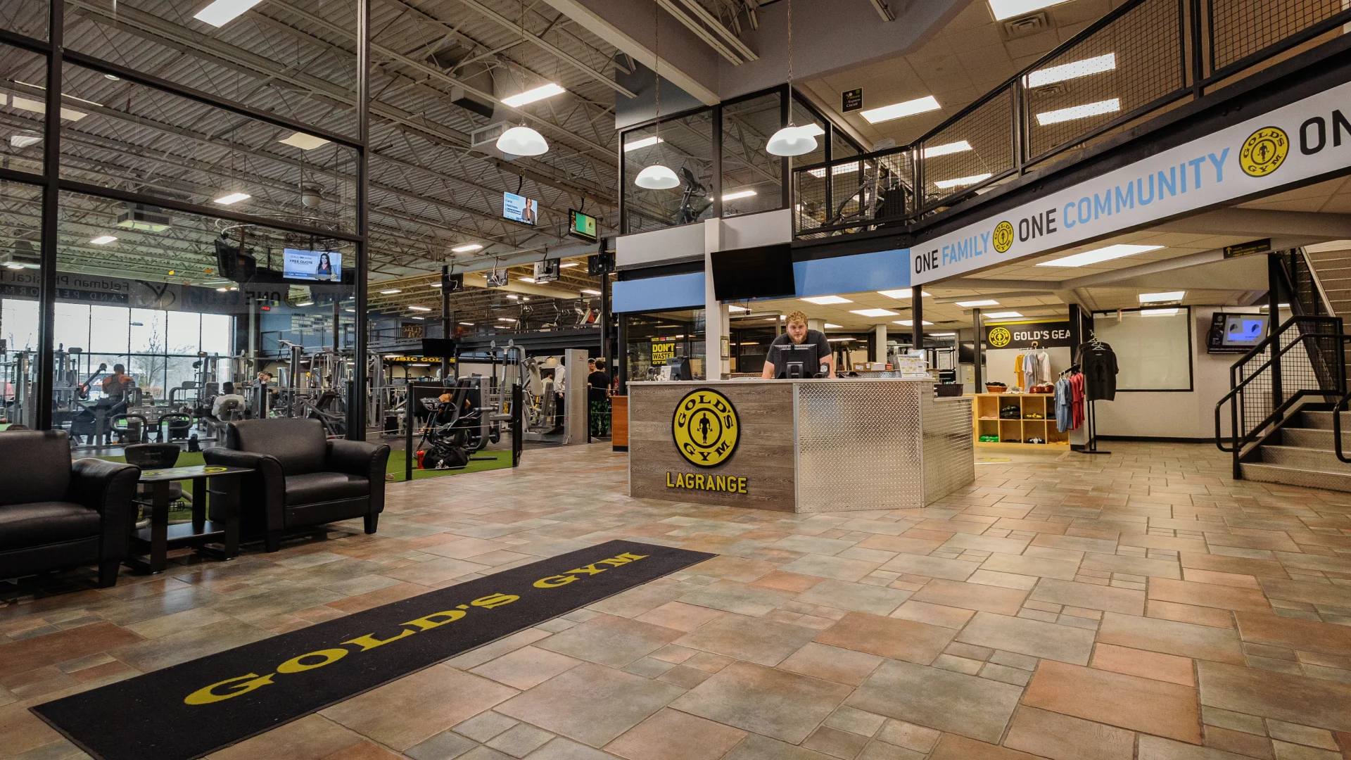 Front desk at Gold's Gym New York (Poughkeepsie), featuring a modern reception area with a wood and metal-paneled counter displaying the Gold’s Gym LaGrange logo. Pendant lights hang from the high ceiling, casting a warm glow over the tiled flooring. A lounge area with black leather chairs and a small table sits near large glass windows overlooking the gym floor, filled with strength equipment. A bold banner above the desk reads “ONE FAMILY, ONE COMMUNITY,” emphasizing the gym’s welcoming atmosphere.