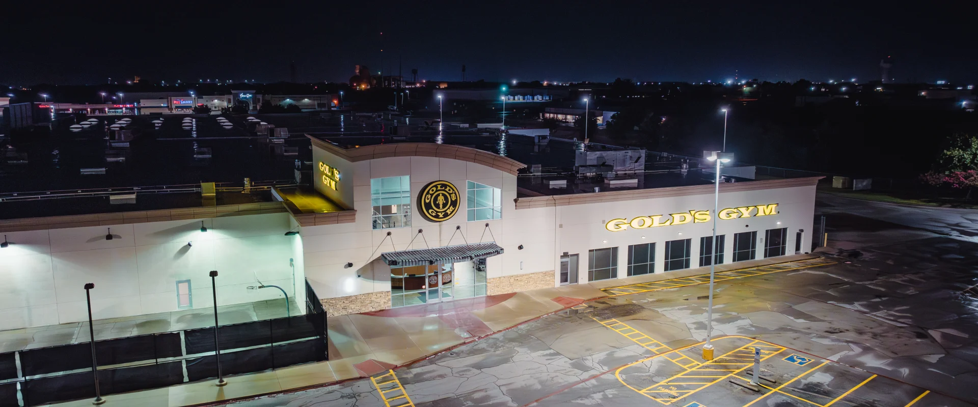 Nighttime exterior of Gold's Gym Waco featuring two illuminated logos, a well-lit entrance with modern architecture, empty parking spaces marked with accessible signage, and surrounding city lights in the background.