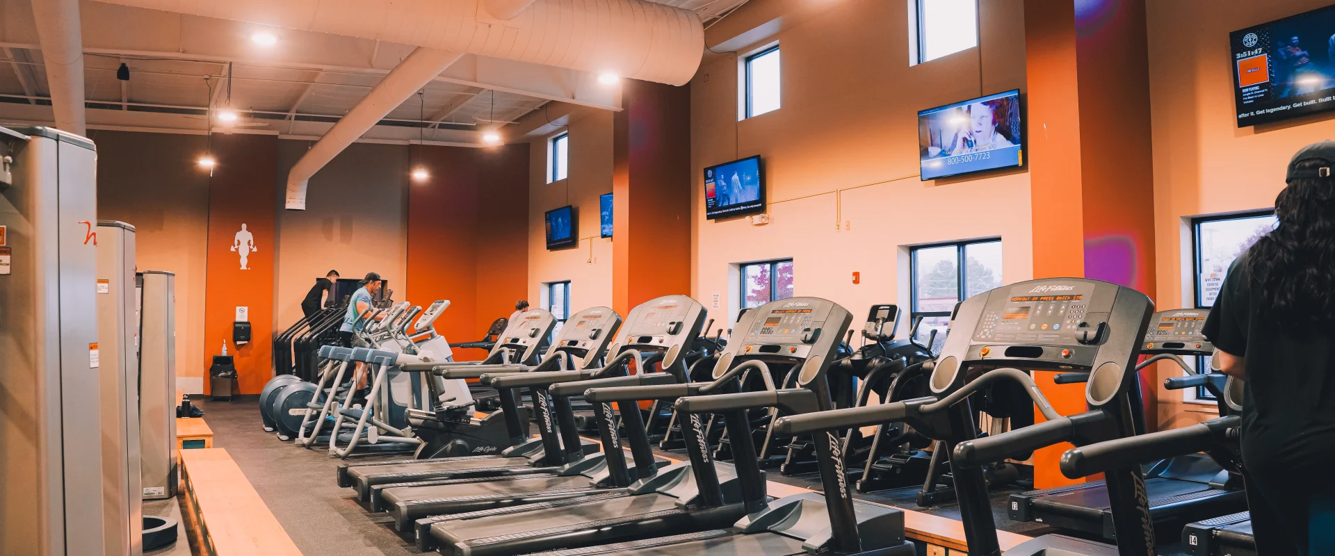 Cardio area at Gold’s Gym Tennessee, Clarksville, featuring a row of treadmills and elliptical machines positioned on a raised platform with a dark rubber floor. The space has high ceilings with large white exposed ductwork, pendant lighting, and a mix of warm-toned walls with windows allowing natural light. Multiple wall-mounted TVs are visible in the background, providing entertainment for gym members during workouts.