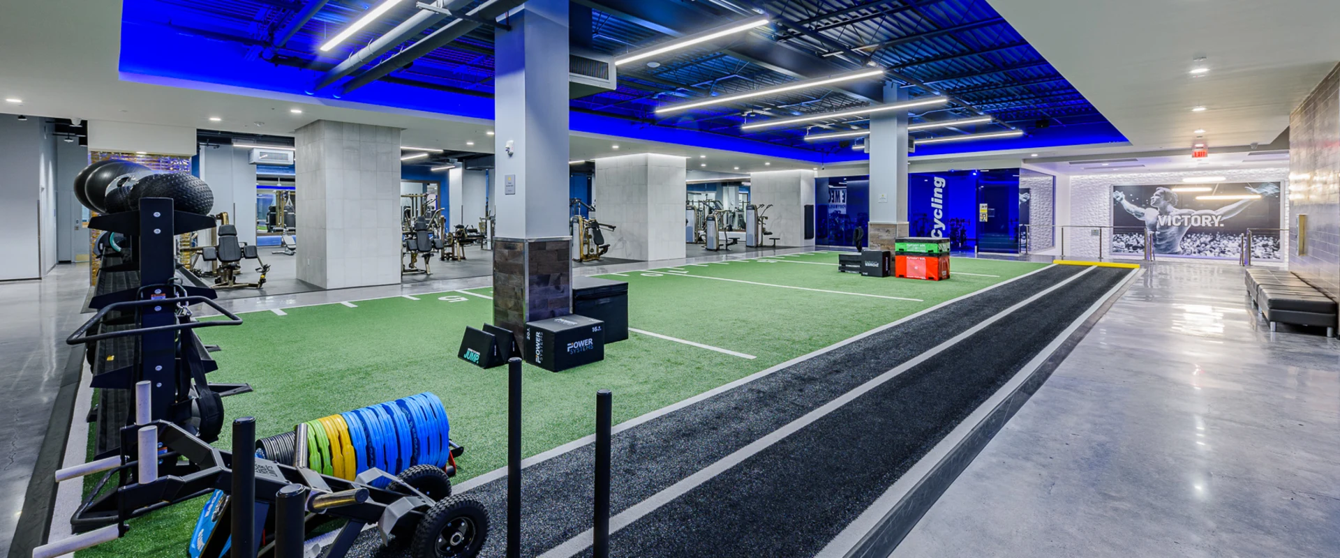 Turf training area at Gold’s Gym Thousand Oaks, designed for functional and athletic workouts. The space features a large artificial turf section with yard line markers, alongside a dedicated sled track. Various training equipment, including weighted sleds, battle ropes, resistance bands, and plyometric boxes, is organized throughout the area. Overhead lighting and an open floor plan create an energetic and spacious environment, seamlessly connecting to the strength and cardio sections of the gym.