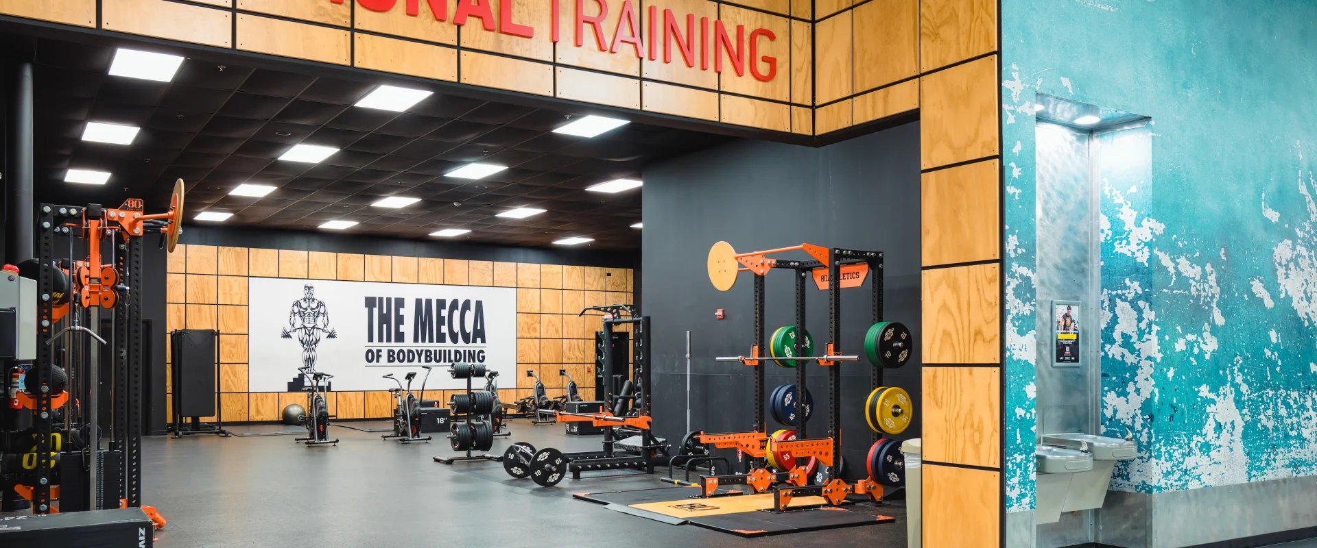 Gold's Gym Crossraods with "Functional Training" sign. Equipment includes weights and racks. Wall reads "The Mecca of Bodybuilding." Energetic vibe.