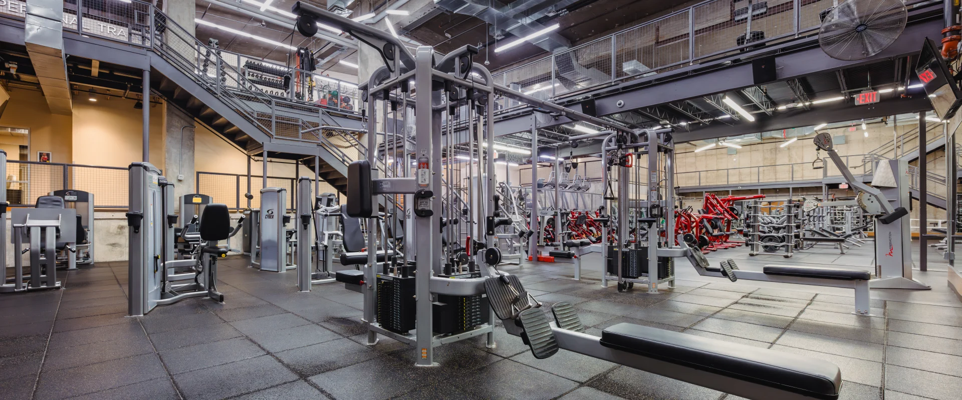 Machine weight area at Gold's Gym Austin (Downtown) featuring strength training equipment on a black rubber floor, with industrial-style lighting and exposed ceilings. A mezzanine staircase leading to an upper-level workout area visible in the background.