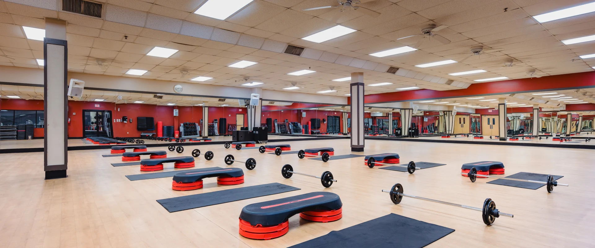 Spacious gym with wooden floors, red and black step platforms, mats, and barbells evenly arranged for a structured workout class. Bright lighting, mirrors along walls.