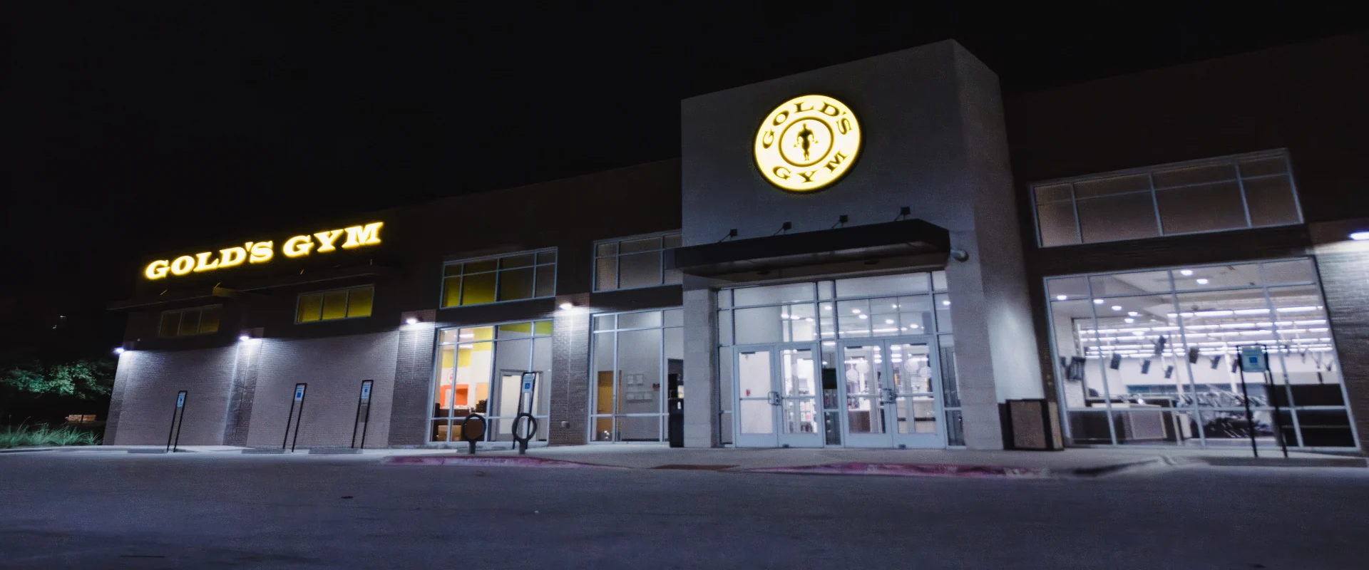 Exterior of Gold’s Gym in Texas (N. Round Rock), showcasing a modern facade with large glass windows and an illuminated Gold’s Gym logo above the main entrance. The bold yellow “GOLD’S GYM” lettering is displayed on the left side of the building. Exterior lights line the building, and the parking area includes accessible spaces with bike racks near the entrance. Inside, cardio equipment and TV screens are visible through the windows.