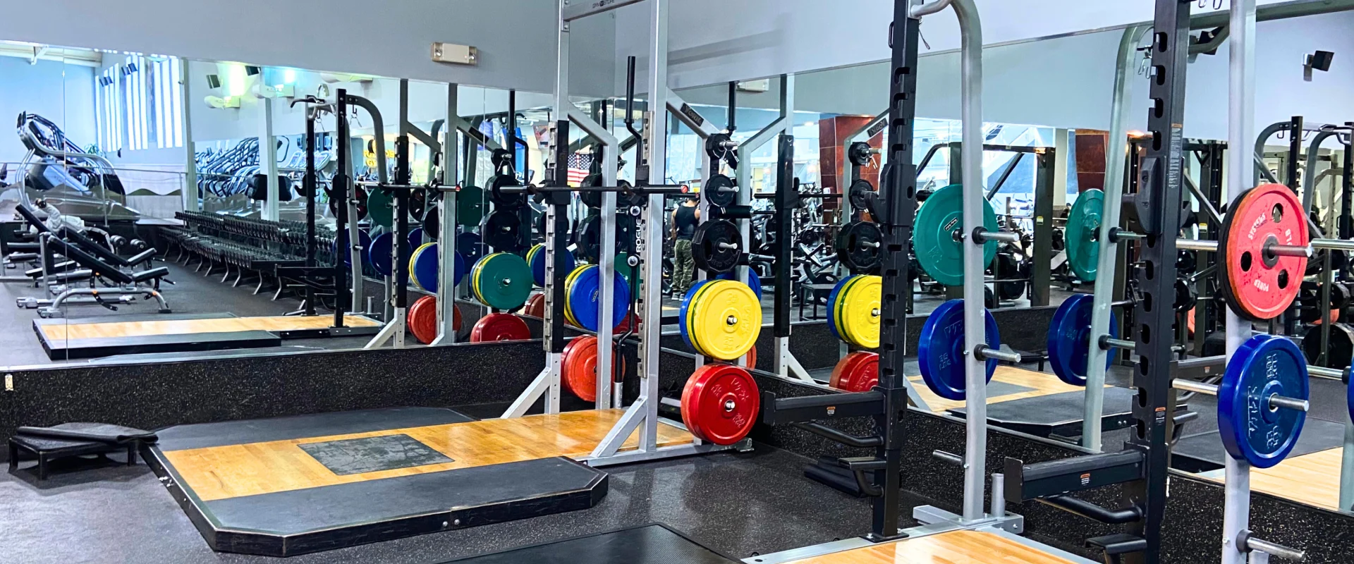 Olympic lifting platforms with power racks in a gym featuring a variety of brightly colored weight plates organized on racks. The platforms have wooden centers surrounded by rubber for shock absorption, with large mirrors lining the wall to reflect the equipment and gym space. Adjustable benches and rows of dumbbells are visible in the background, creating a comprehensive strength training area.