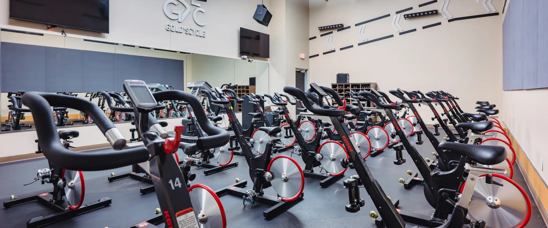A spacious indoor cycling studio features rows of stationary bikes with red accents on a dark floor. Mirrors and large screens adorn the walls, creating an energetic ambiance.