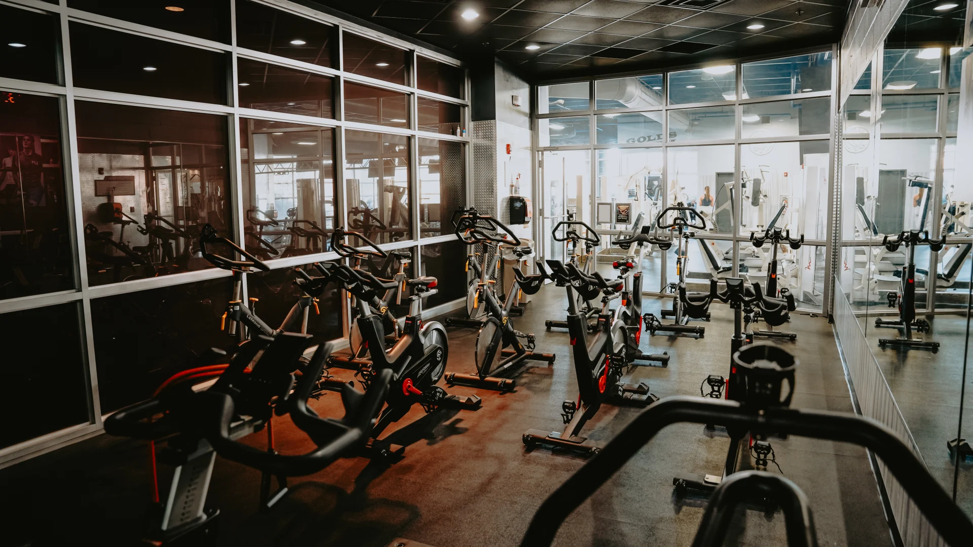 Group cycle room at Gold’s Gym Tennessee, Downtown Chattanooga, featuring rows of stationary bikes on a dark rubber floor. The space includes a mirrored wall, glass partitions, and overhead lighting. A hallway and adjacent workout area with weight equipment are visible through the glass, creating a bright and open atmosphere.
