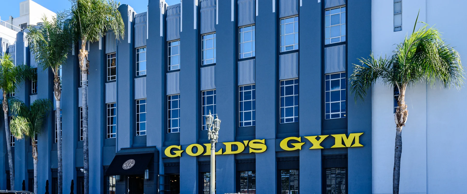 Exterior of Gold’s Gym Hollywood, featuring a multi-story gray building with tall, vertical architectural details. The gym's name is displayed in large, bold yellow letters above the entrance. A black awning with the Gold’s Gym logo marks the main entrance. Several parked cars line the street, and palm trees frame the building. A decorative street lamp stands in front of the entrance.
