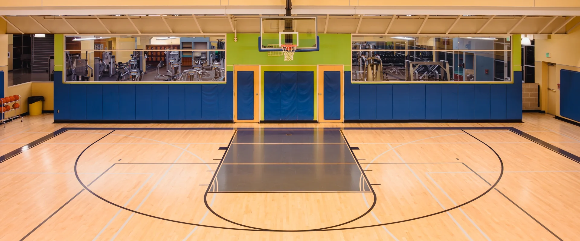 Empty indoor basketball court with polished wood flooring and hoop. Behind, large glass windows reveal a fitness area with gym equipment. Bright, spacious setting.