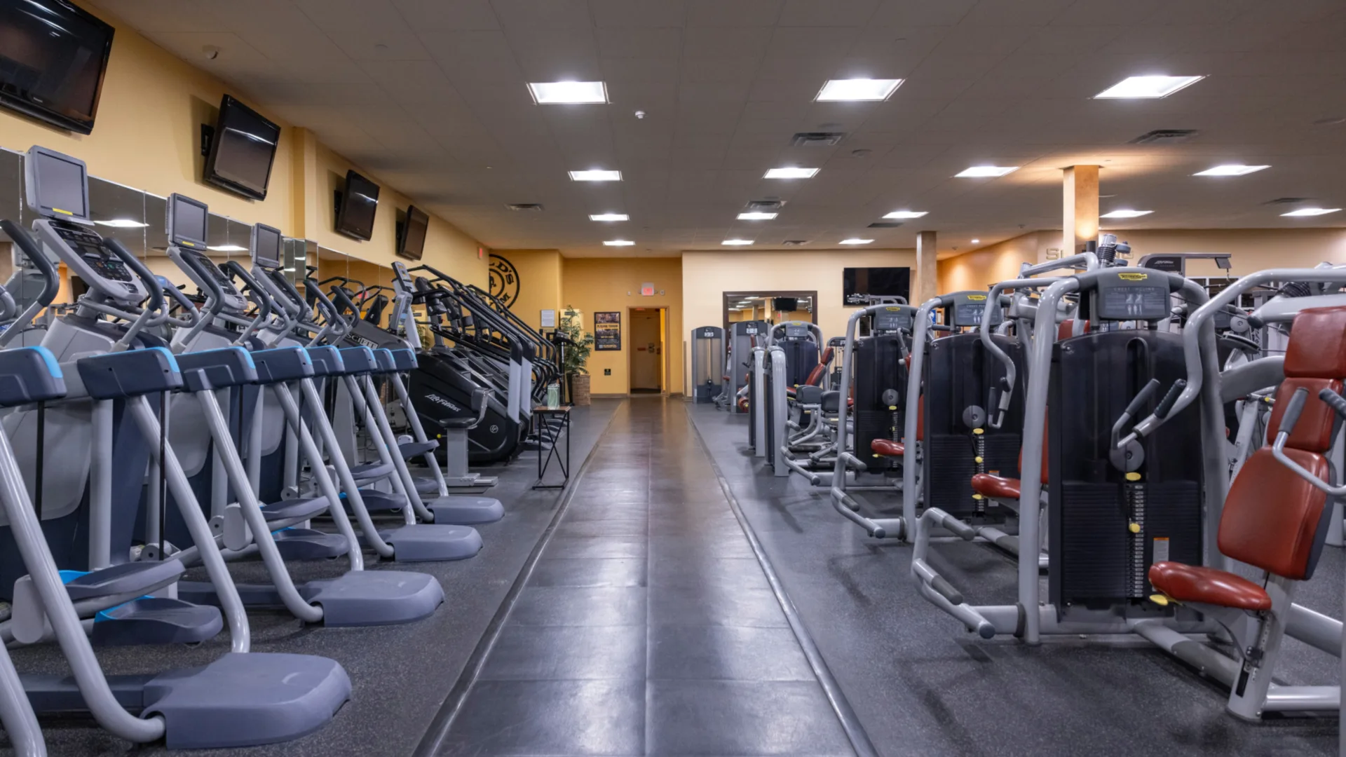 Strength equipment area at Gold’s Gym in East Northport, New York, featuring Life Fitness selectorized machines with padded red seats for targeted muscle training. The space includes rows of treadmills and stair climbers along mirrored walls with mounted TVs overhead. Bright ceiling lights and rubber flooring create a clean, organized workout environment.
