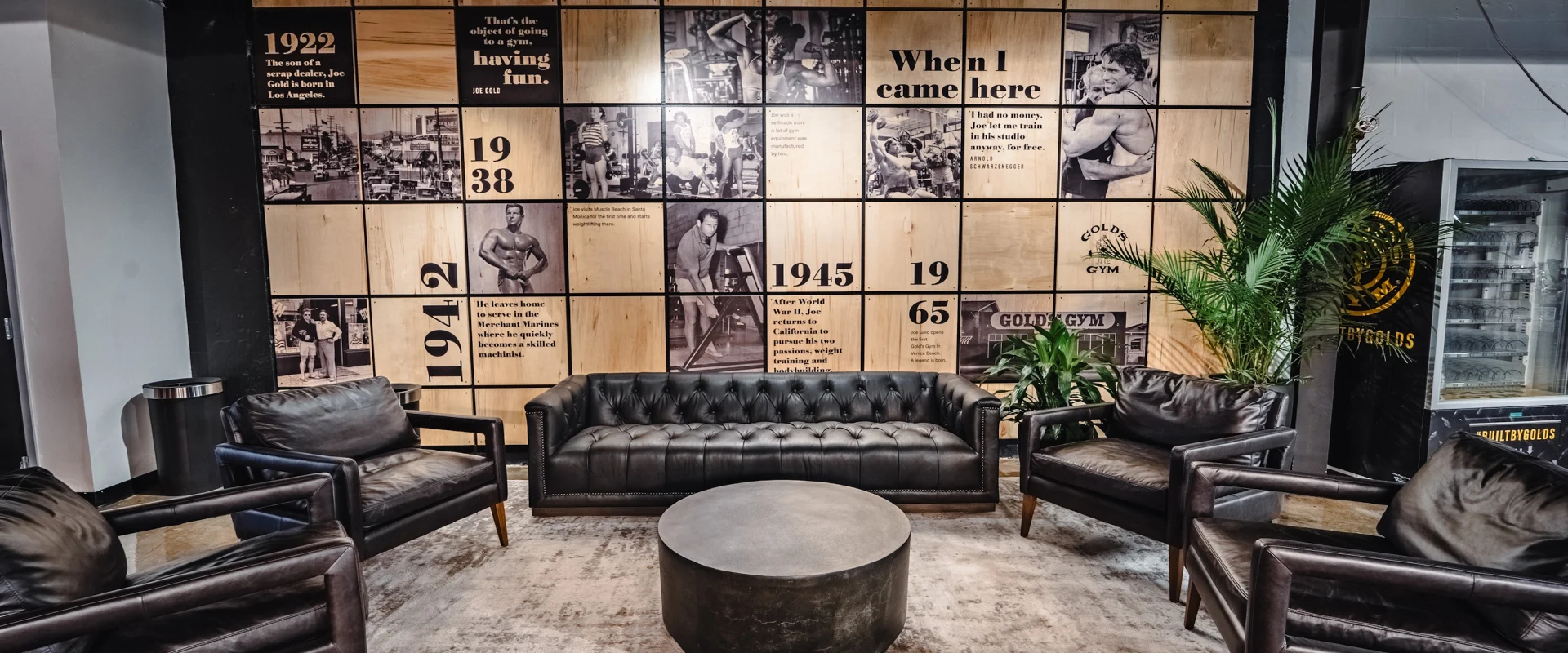 Lobby seating area at Gold’s Gym (Gas Worx) with leather chairs and a wall mural featuring Gold’s Gym history.