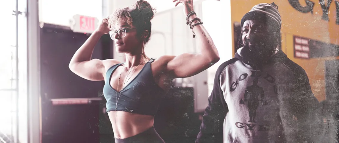 The image depicts two muscular individuals, one in the foreground wearing a sports bra and the other in the background wearing a dark outfit, both with their arms raised in a gym or fitness setting.