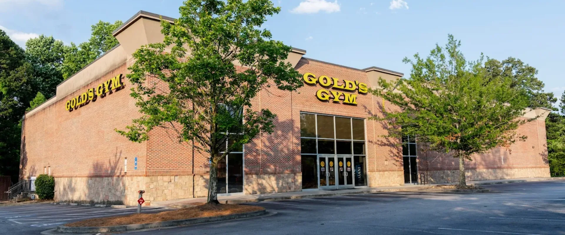 Exterior of Gold’s Gym Hamilton Mill with yellow signage on brick building and glass entry under clear blue sky.