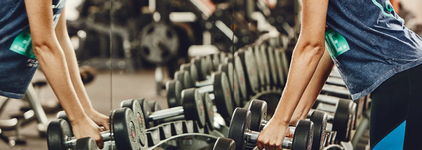 A collection of various weights and dumbbells arranged in a gym setting, with a person's arm visible in the foreground.