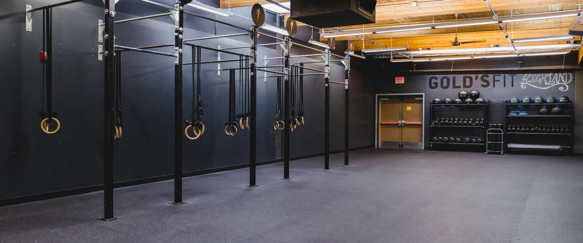 A Gold's Fit training area with gymnastic rings, pull-up bars, medicine balls, kettlebells, and a sleek black interior under a wooden ceiling.