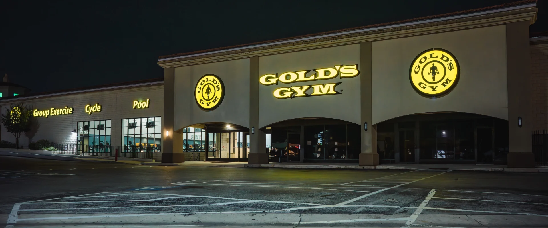 Exterior of a Gold's Gym at night, brightly lit with glowing yellow signs. The facade includes group exercise, cycle, and pool areas, exuding an active vibe.