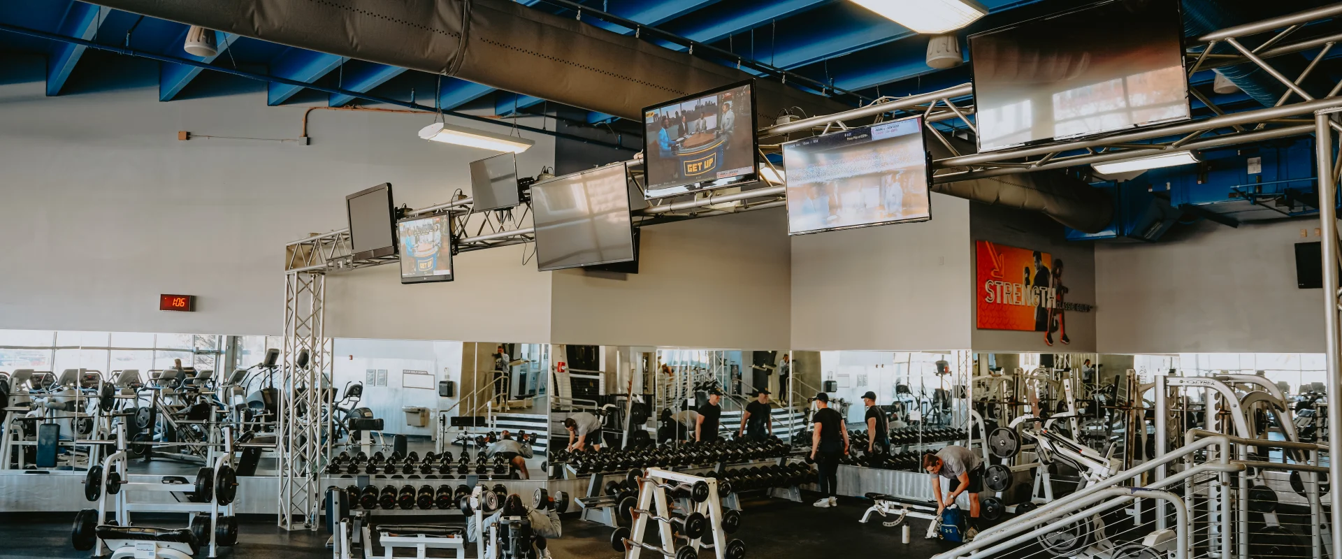 Dumbbell area at Gold’s Gym Tennessee, Downtown Chattanooga, featuring a neatly arranged row of dumbbells on racks against mirrored walls, creating an expansive feel. The space includes a dark rubber floor, high ceiling with exposed blue ducts, and overhead lighting. Mounted televisions and a motivational “Strength” sign enhance the workout environment, with reflections of the gym space visible in the mirrors.