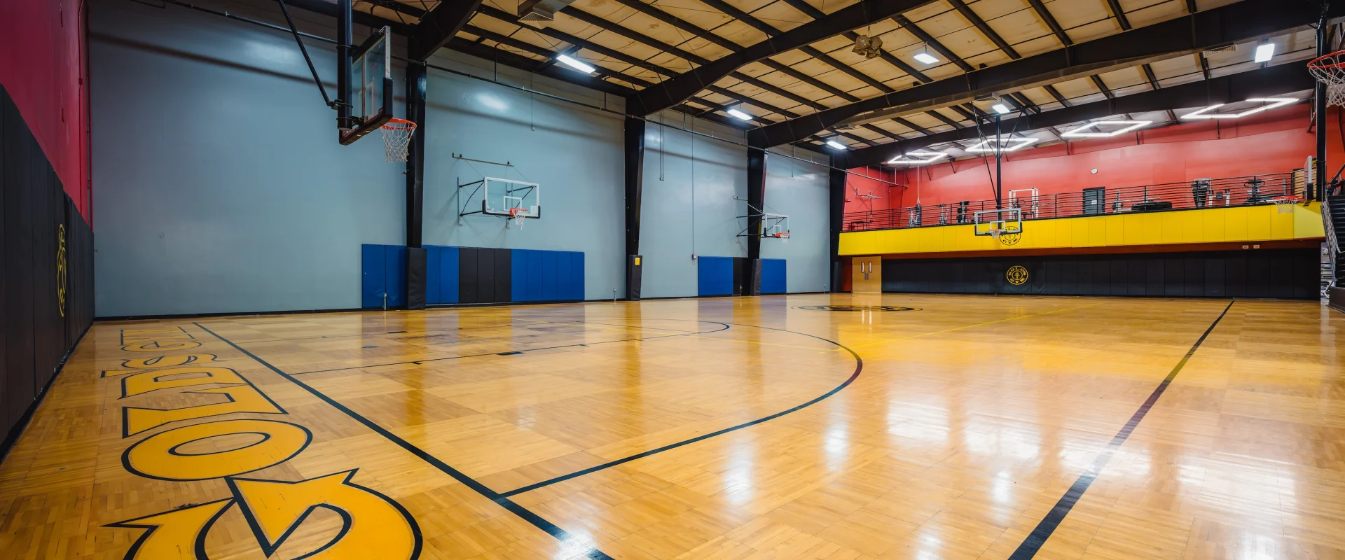 Indoor basketball court at Gold's Gym Babcock with polished wood flooring, red and blue walls, multiple hoops, and bright lighting. Emphasizes readiness and energy.
