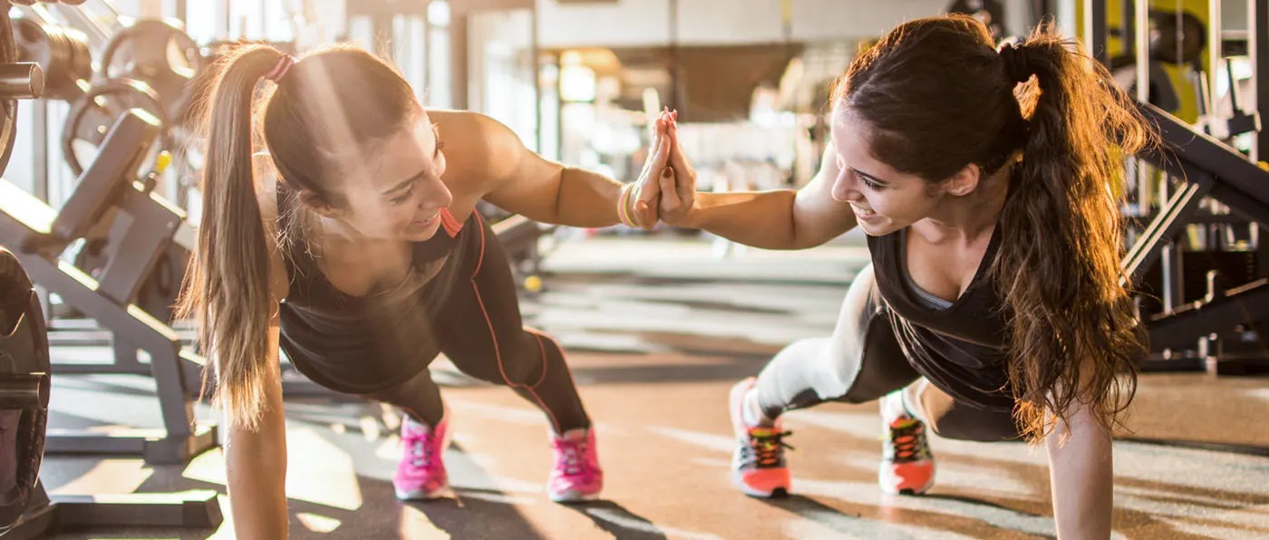 Two young women in athletic wear are performing push-ups together in a well-equipped gym, with various exercise equipment visible in the background.