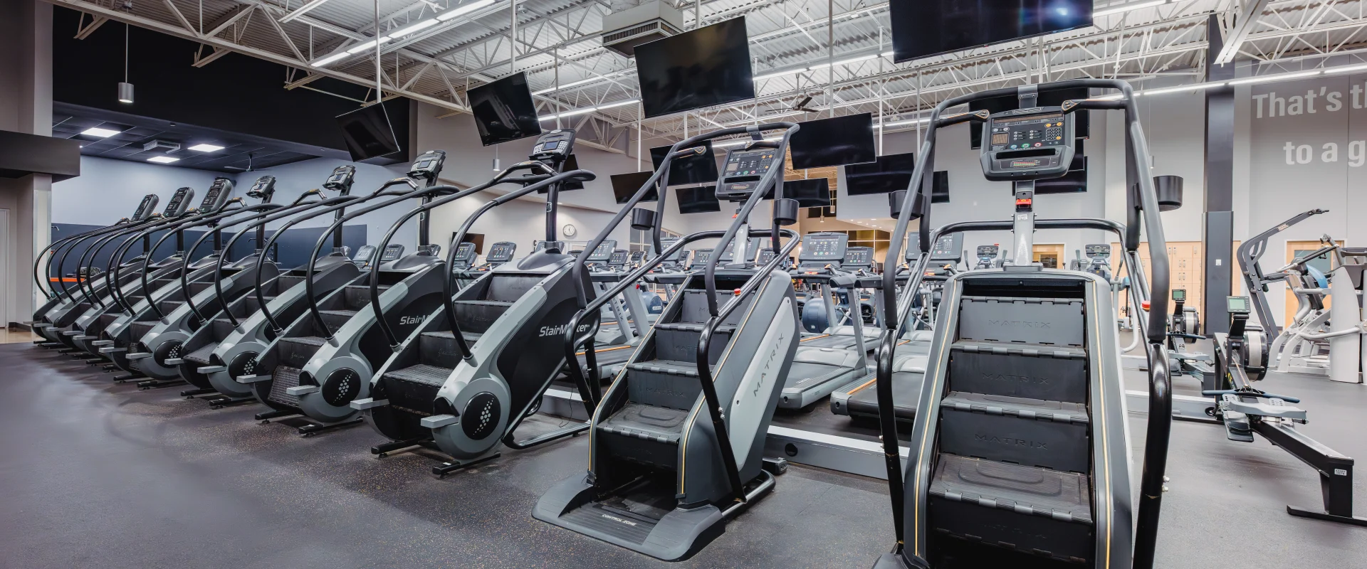 A cardio area at Gold's Gym featuring rows of stair climbers and treadmills under a high ceiling with bright lighting and multiple mounted TVs.