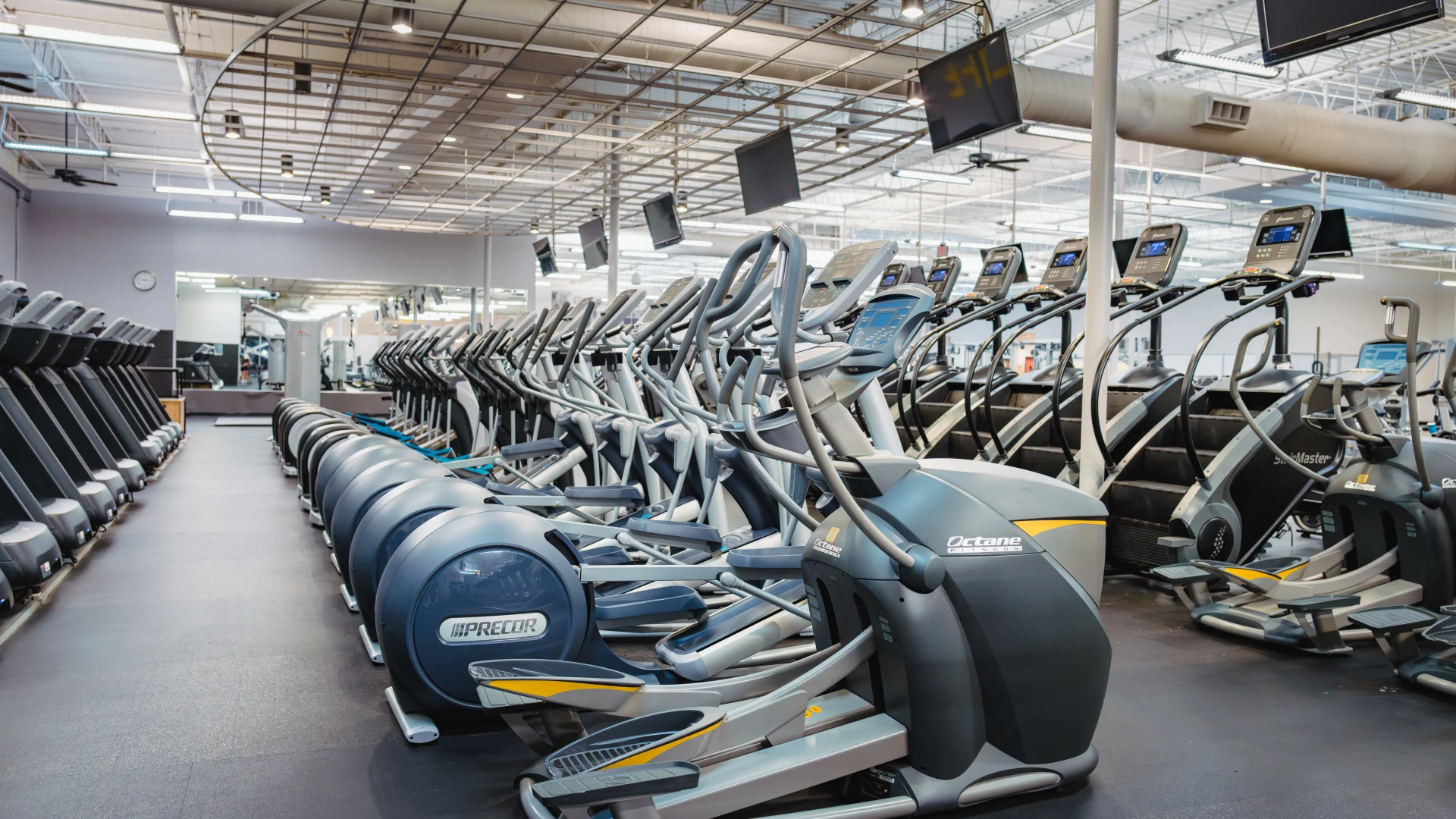 Cardio area at Gold's Gym in Austin (Hesters Crossing) featuring rows of elliptical machines and treadmills aligned on a sleek black floor. The space is brightly lit with overhead industrial-style lighting and features mirrors along the back wall to enhance the open feel. Mounted TVs hang from the ceiling, offering entertainment for gym members during workouts.