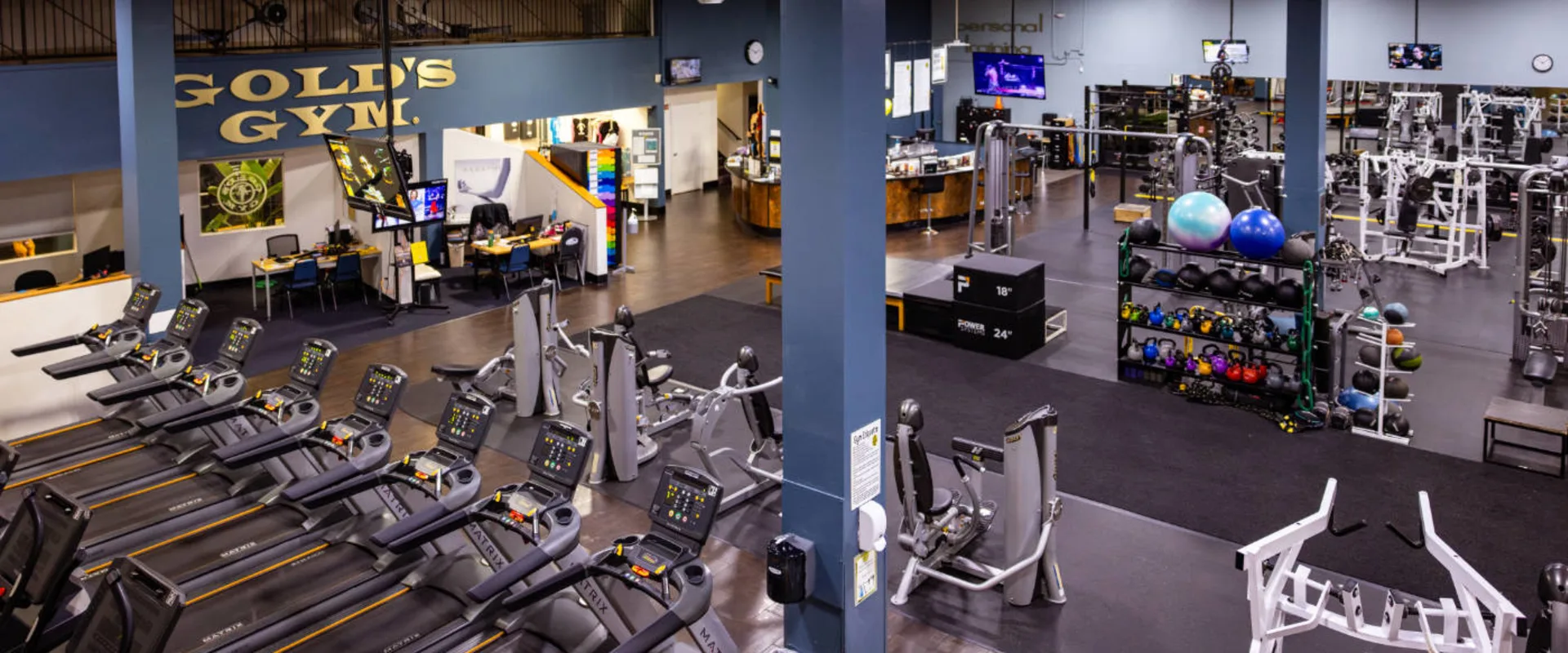 An elevated view inside Gold’s Gym in Woodbridge (Lake Ridge) shows a spacious layout with various workout zones. In the foreground, a row of Matrix treadmills lines the black rubber flooring. To the right, strength training machines and free weights are organized near a functional training area equipped with kettlebells, medicine balls, and plyometric boxes. The gym’s blue structural columns contrast against the warm-toned wood flooring near the front desk and retail area. The Gold’s Gym logo is prominently displayed on the back wall, reinforcing the brand’s presence. Multiple wall-mounted TVs and digital screens are visible throughout the space.