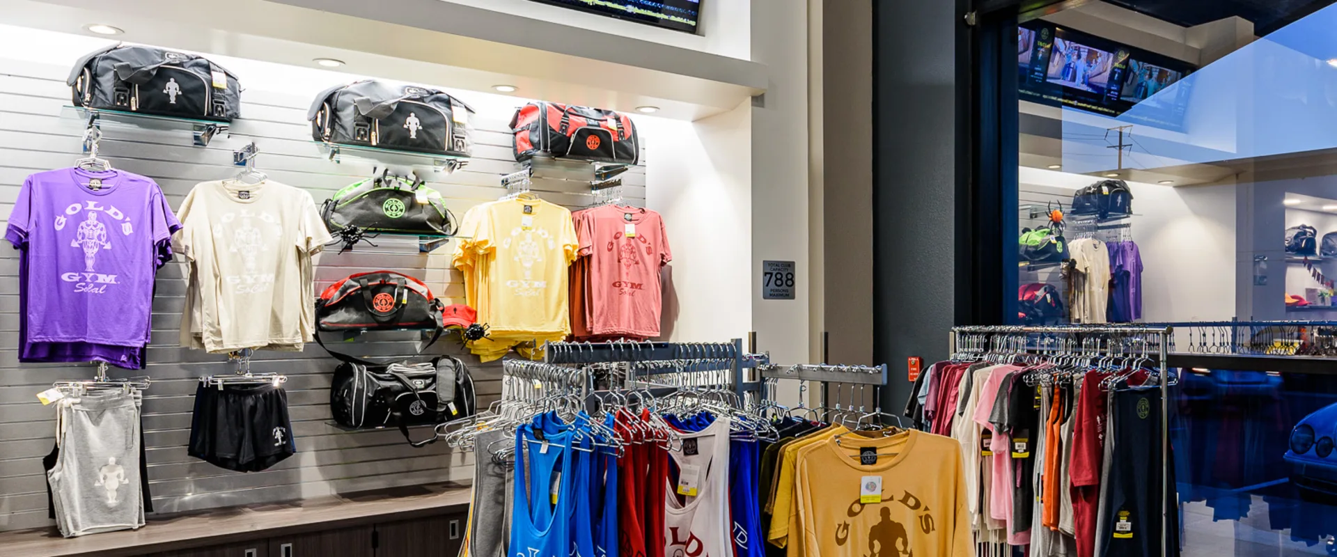 A well-stocked Gold’s Gym Pro Shop showcasing branded apparel, including t-shirts, tank tops, gym shorts, and bags neatly displayed on racks and shelves. Multiple digital screens above the merchandise display promotional gym content, while floor-to-ceiling windows allow natural light into the retail space.