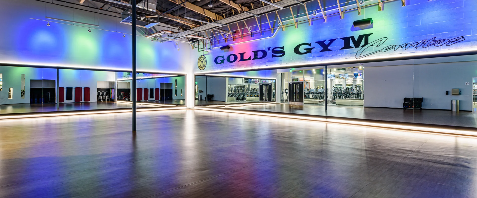 The group exercise studio at Gold’s Gym Cerritos features a spacious wood-floored area with a mirrored wall running its length. Soft LED lighting casts blue and green hues onto the high ceiling, which includes exposed ductwork and industrial fixtures. A "Gold’s Gym Cerritos" sign is displayed on the wall above the mirrors. The studio is equipped with wall-mounted speakers and storage racks for workout accessories.