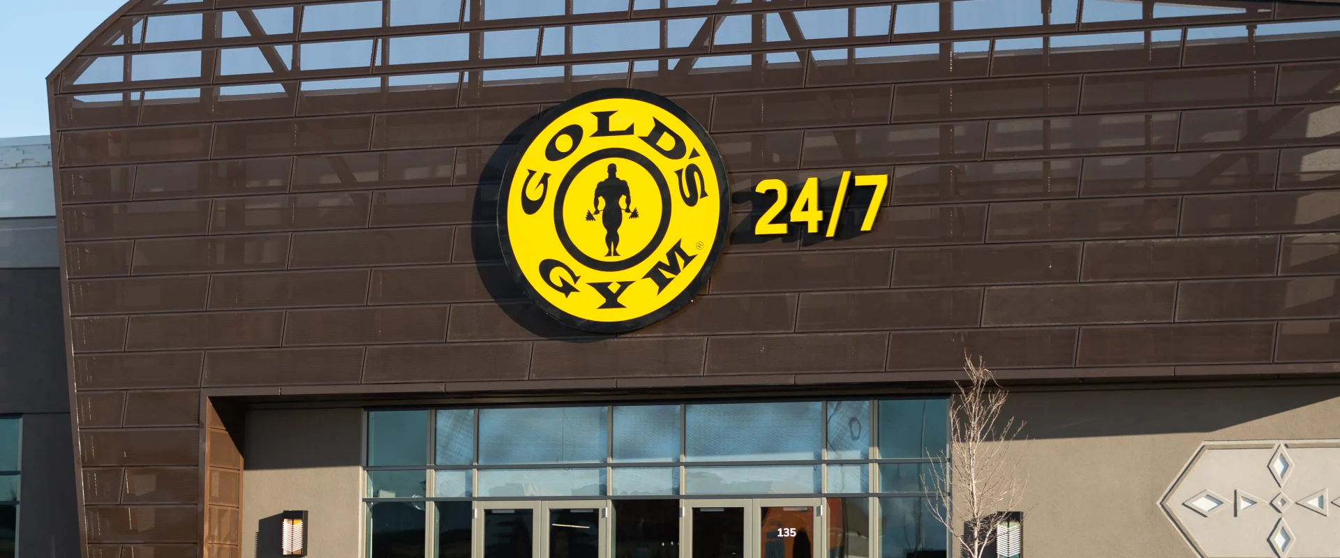 Exterior of Gold’s Gym Canada, Buffalo Run, Alberta, showcasing a modern architectural design with a distinctive curved roof structure and large glass windows. The front entrance features the iconic yellow and black Gold’s Gym logo accompanied by "24/7" signage, highlighting the gym's accessibility. The surrounding area includes a neatly maintained walkway, light snow coverage, and a bench near the entrance.