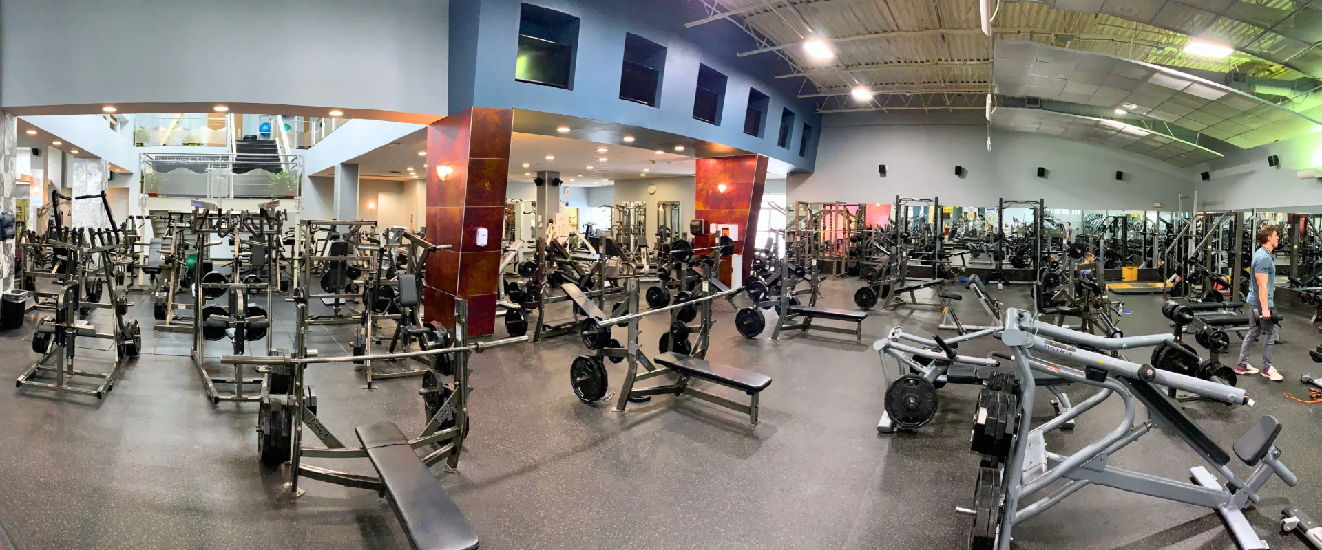 Spacious free weights area in a Smithtown gym featuring rows of weight benches, squat racks, and plate-loaded machines. The room has high ceilings, bright lighting, and a modern design. Mirrors line the walls, reflecting equipment and gymgoers. A person is using free weights on the right side of the image.