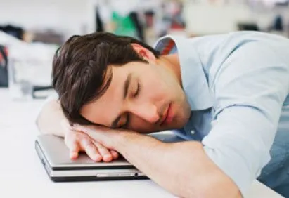 A young man is sleeping at his desk, his head resting on his arms as he appears to have fallen asleep while working on his laptop.