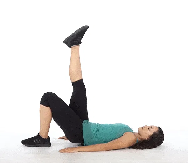A person in black athletic clothing is lying on the floor with their legs raised, appearing to be performing a leg exercise or stretch.