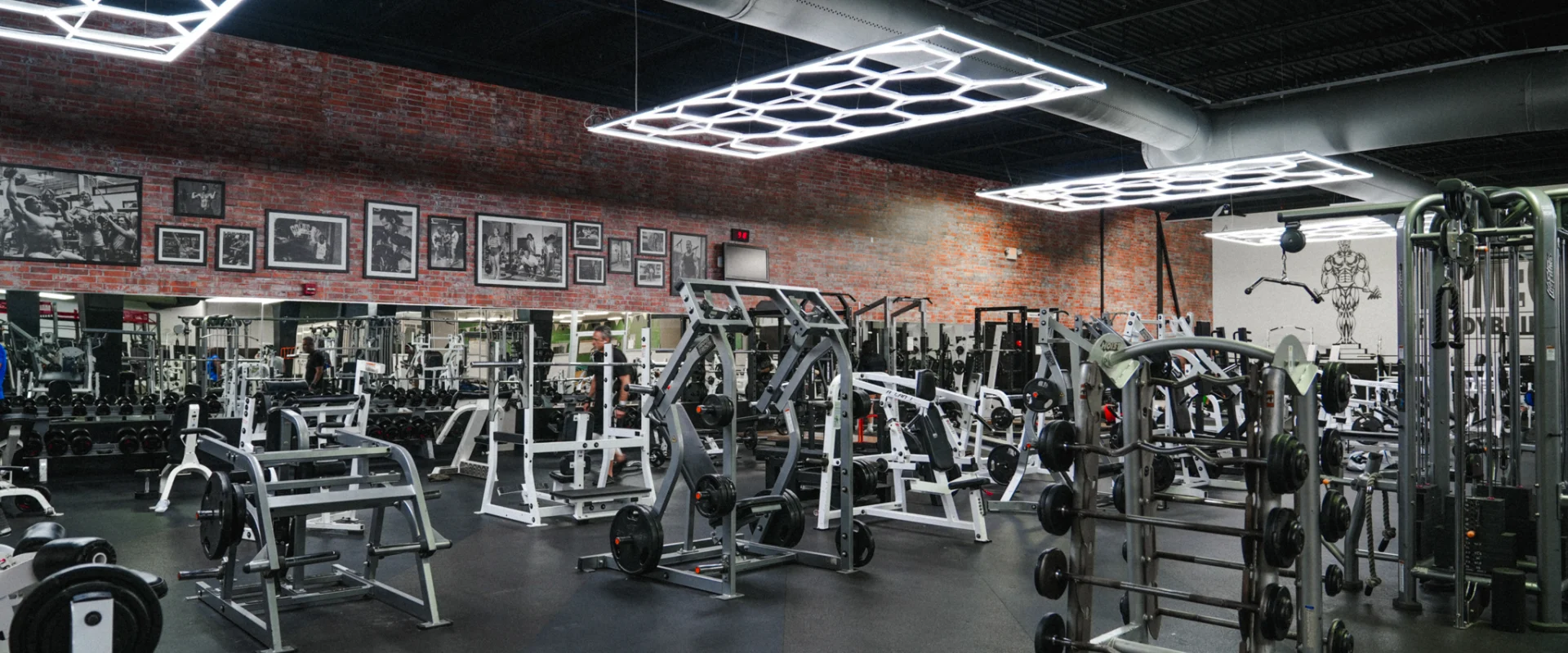 A wide view of Gold's Gym Brassfield's main workout area, filled with plate-loaded and selectorized weight machines. Neon LED lights illuminate the open space, and a red brick accent wall features classic bodybuilding images. The "Mecca of Bodybuilding" mural stands prominently on the far wall.