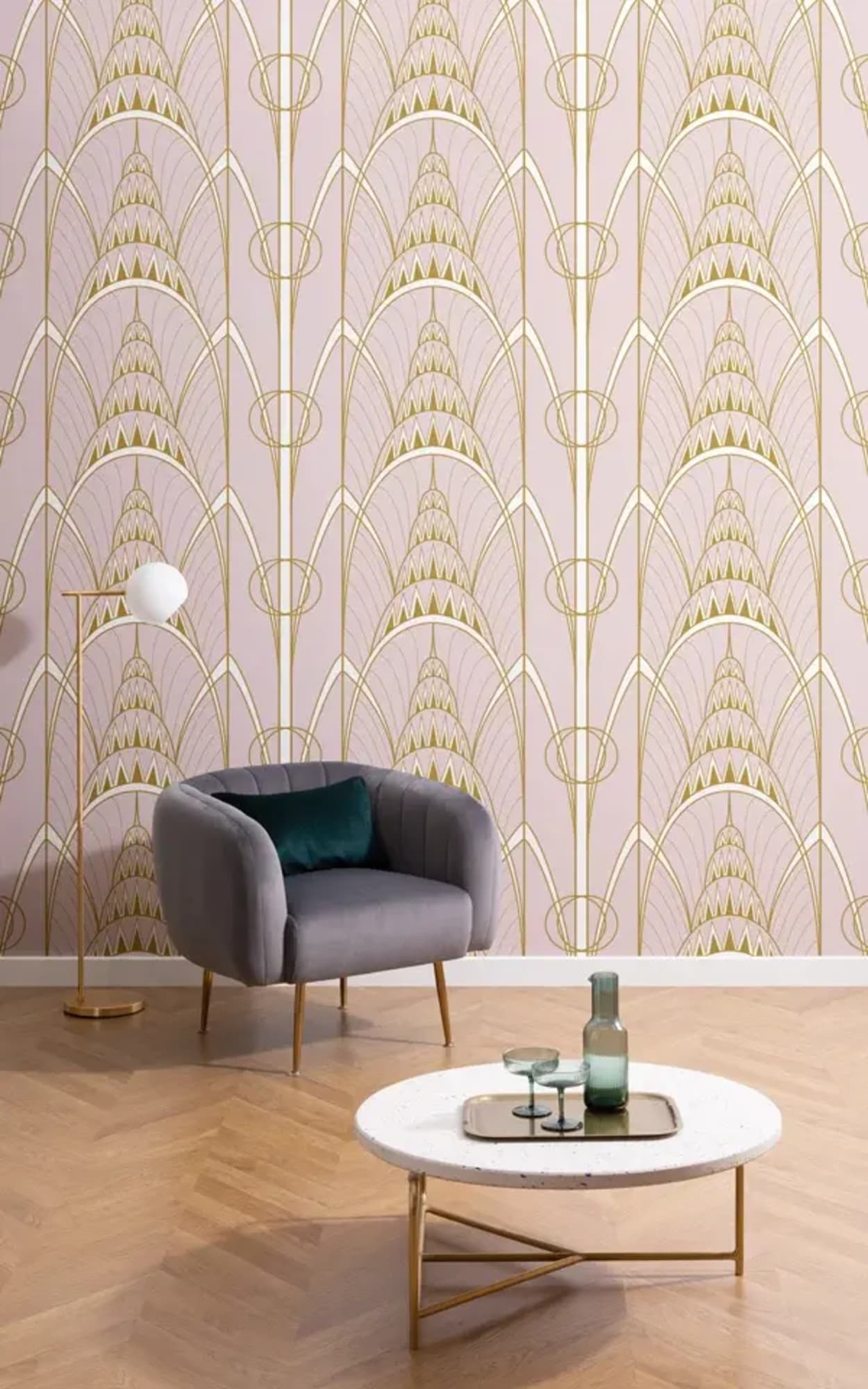 A stylish living room featuring a gray armchair with a teal cushion, a white round coffee table with green glass decor, set against a wall with a pink and gold Art Deco-style wallpaper pattern.