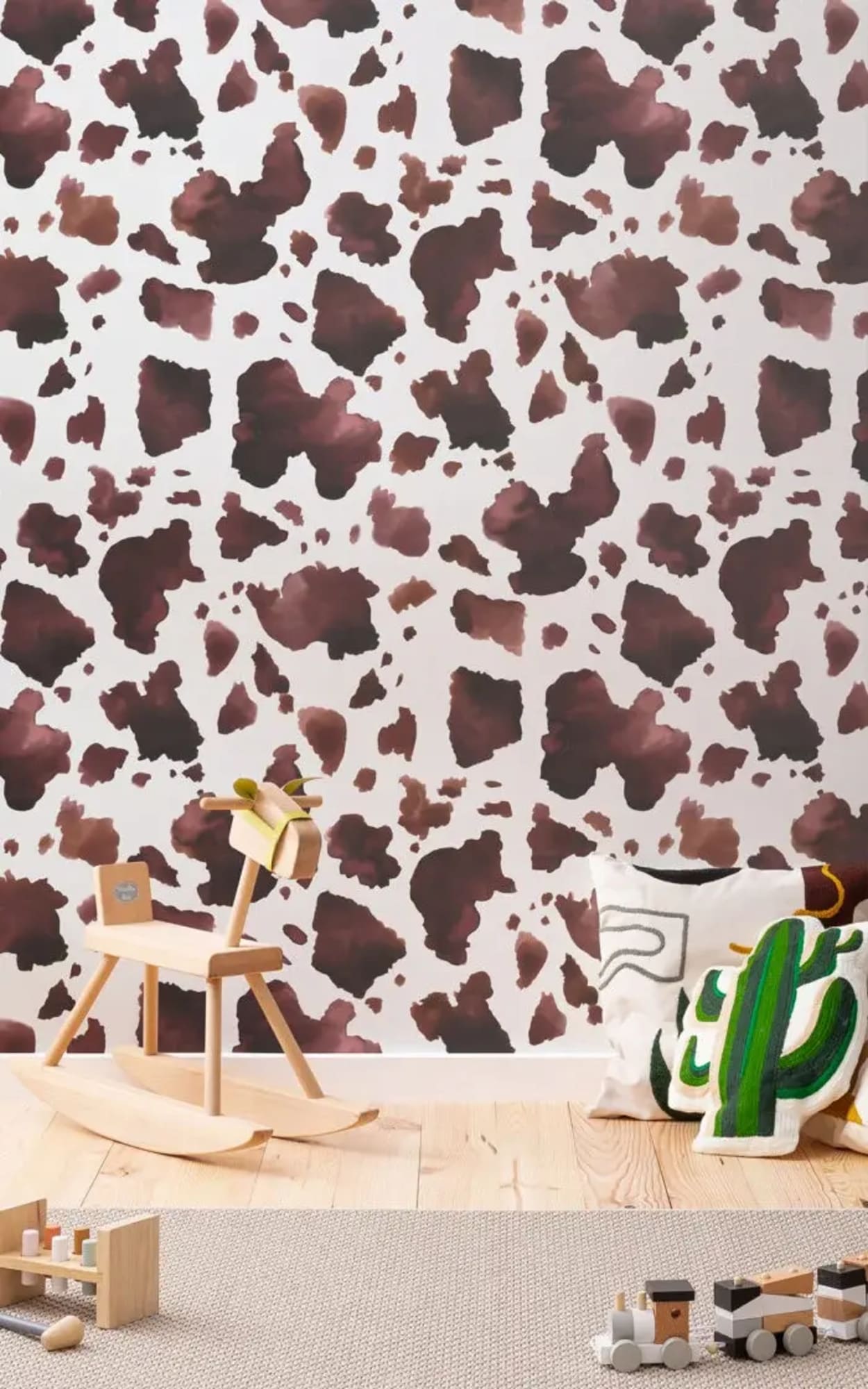 A children's room features a wooden rocking horse, decorative pillows, and toys, set against a wall with a brown cow print pattern.