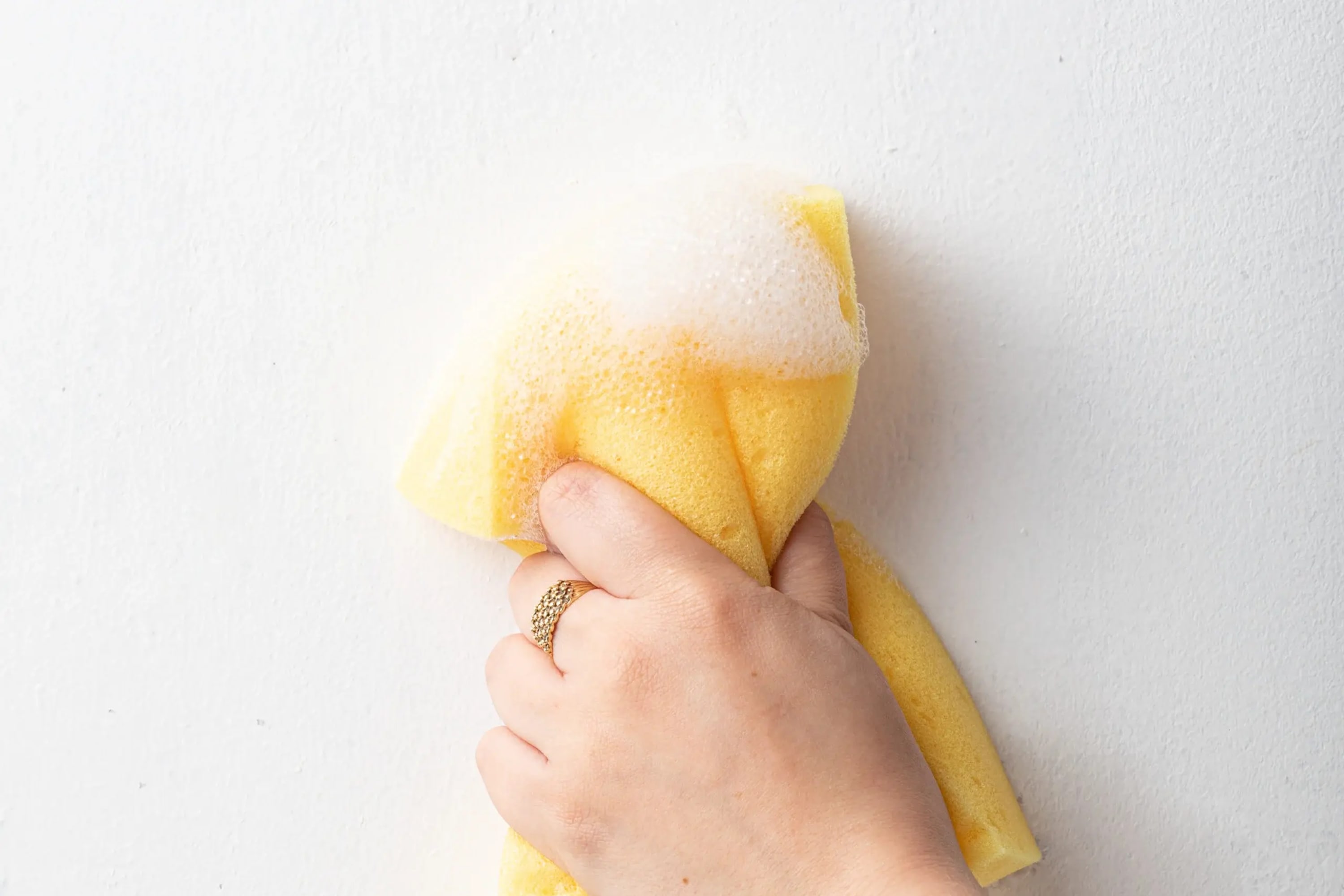 A hand wearing a gold ring cleans a white wall with a yellow sponge and soapy foam.