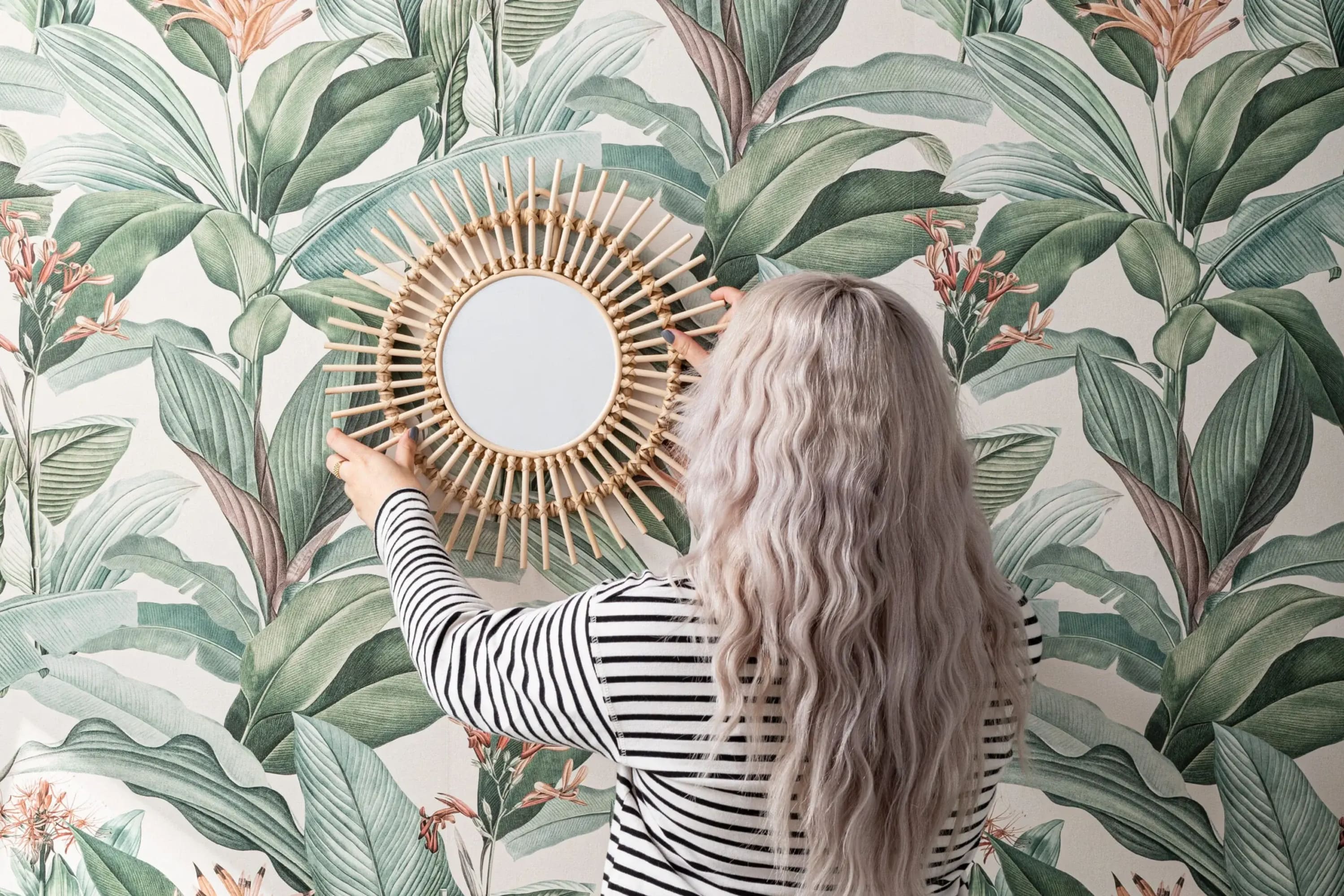 A person hangs a round wicker mirror on a wall with green and pink botanical wallpaper.