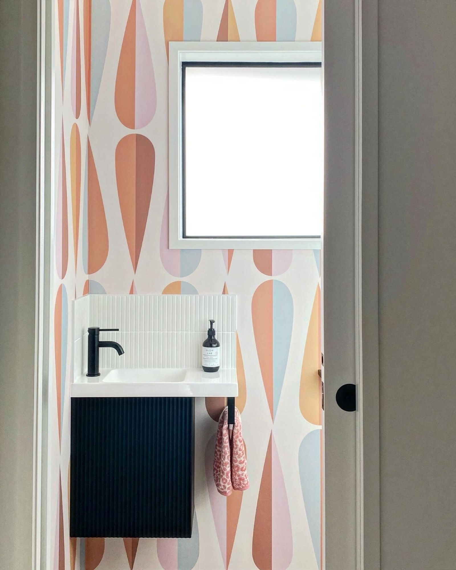 A small bathroom with colorful geometric wallpaper, a window, a white sink with a black faucet, and a black cabinet underneath.