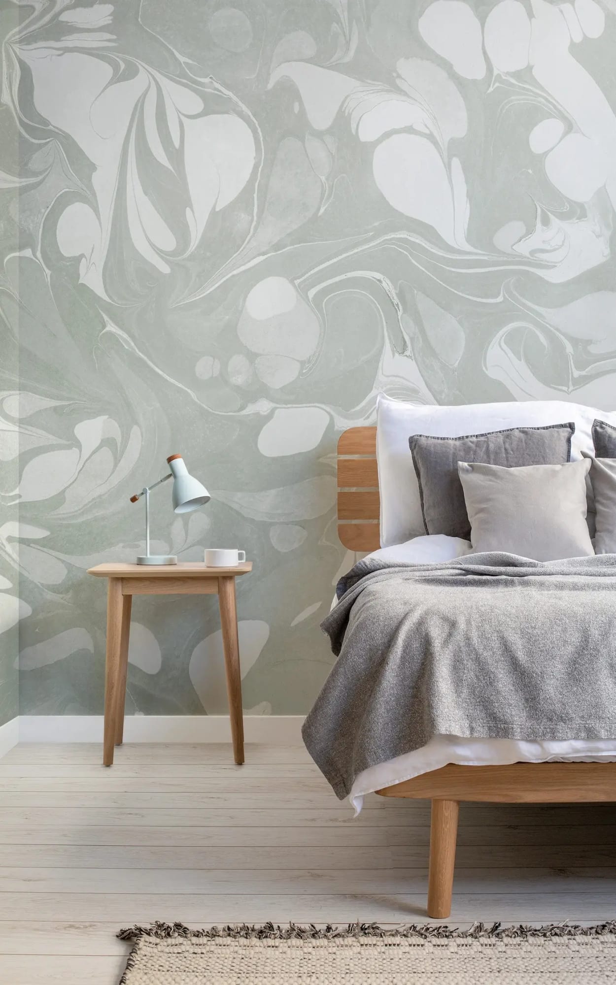 Stylish bedroom with a bed covered in gray bedding, adjacent to a wooden bedside table with a blue desk lamp, set against a large abstract green and white mural.