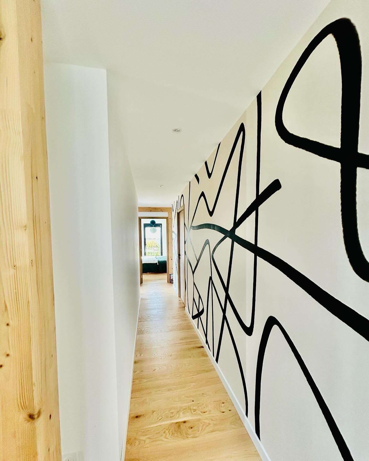 A modern hallway with a bold black abstract line mural on a white wall, light wood flooring, and a door leading to a bright room.