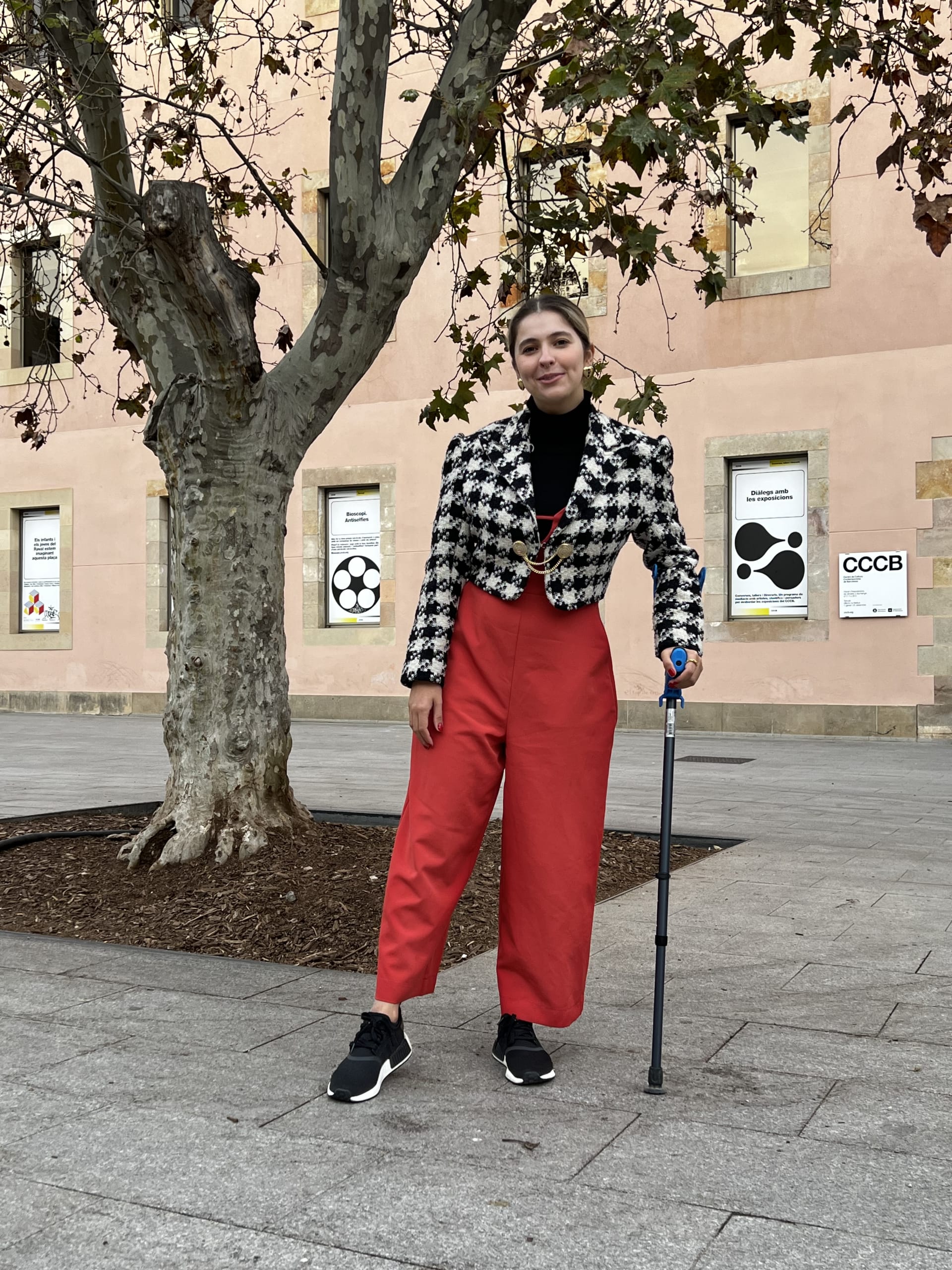 Foto ilustrando looks com macacão. Marcie está em um museu rosa de fundo. O look é composto por blusa de gola alta preta por baixo de macacão laranja e jaqueta cropped preta e branco com estampa pied de coq e tênis de corrida preto.