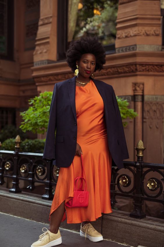 Foto para ilustrar 7 looks para usar essa semana. Uma mulher com cabelo black power preto está em frente a um prédio em tons de marrom. O look é composto por vestido acetinado longo laranja com um blazer preto nos ombros, tênis bege e bolsa vermelha.